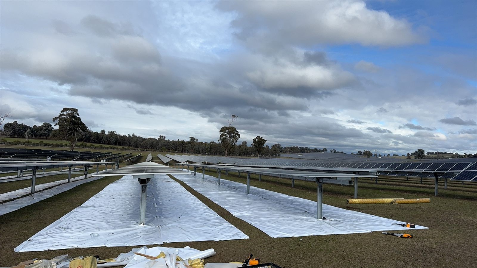 Wide field view of a Sunplus install in Australia: reflective mat laid across tracker rows with gum trees on the horizon and installer tools in the foreground.